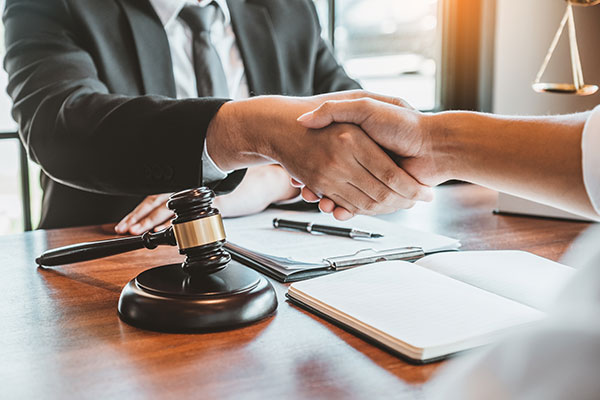 An attorney and client shaking hands over a wooden desk with a gavel, pen, notebook, and clipboard on it