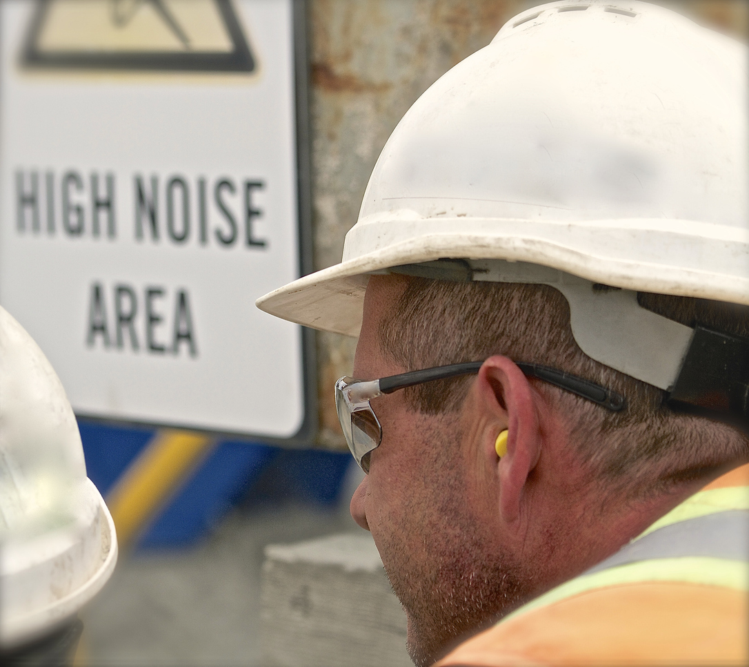 A construction worker with earplugs standing next to a high-noise area sign