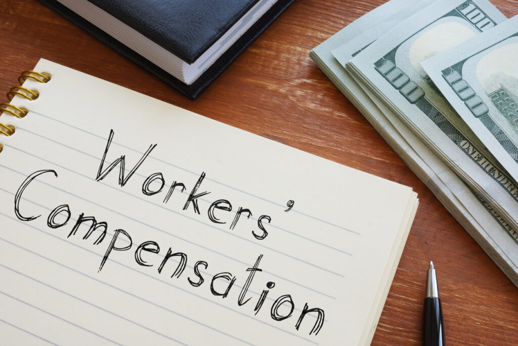 Close-up of a notebook page with “workers’ compensation” written on it with a pen, along with money and a book