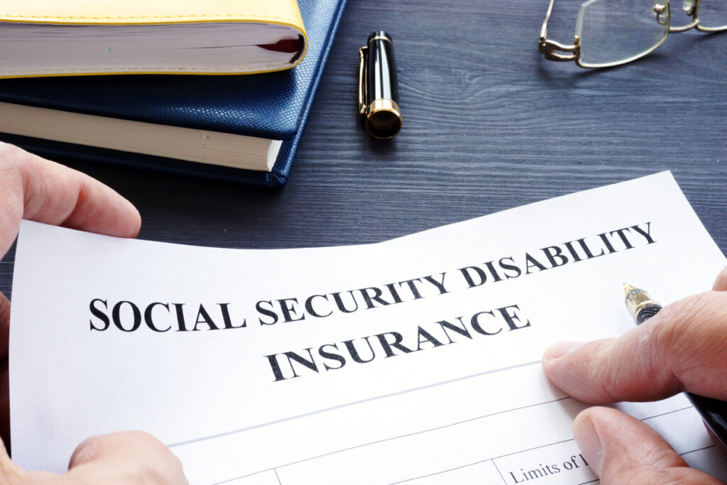 Close-up of a man filling out an SSDI form with books, a pen cap, and eyeglasses in the background