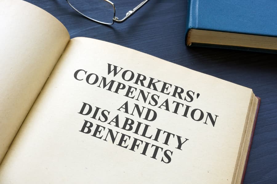 An open book about workers' compensation and disability benefits with a pair of eyeglasses and blue book in the background