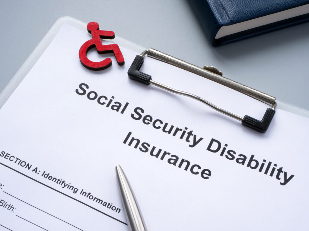 A clipboard with a Social Security Disability form, silver pen, black book, and red disability symbol
