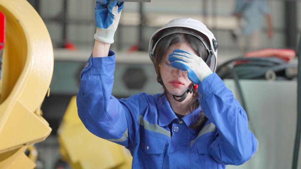 Female worker in industrial setting wearing blue shirt and white hard hat and holding her hand to her face